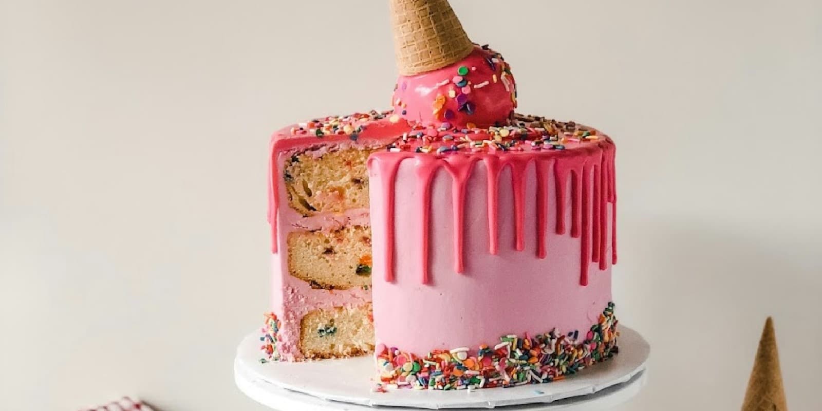 A pink drip cake topped with an ice-cream cone on a white cake stand